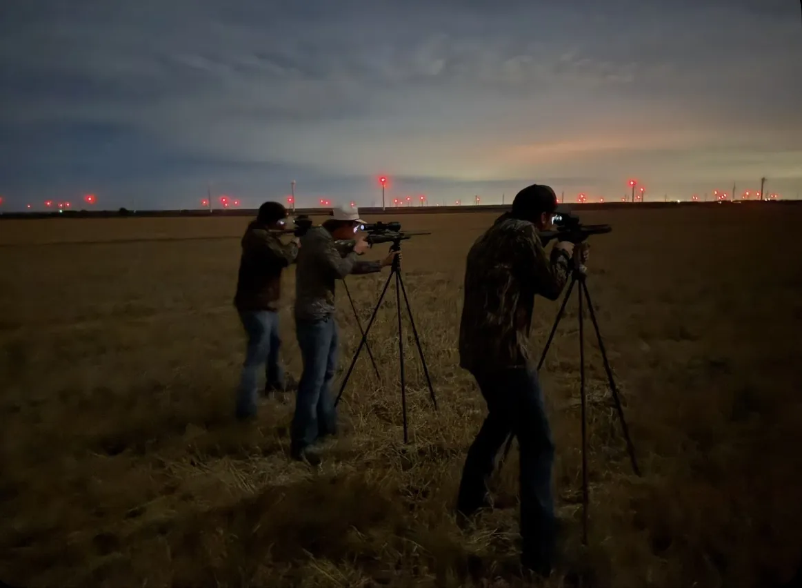 Three hunters aiming tripod-mounted scoped rifles across a West Texas field at dusk, a wind farm on the horizon