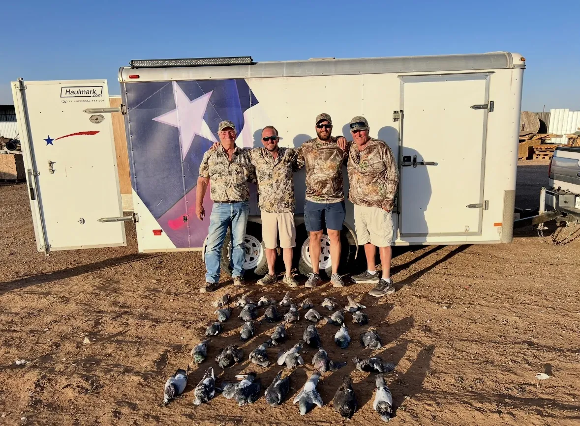 Four hunters standing in front of a white enclosed trailer wrapped with a Texas flag and star, a spread of pigeons fanned across the dirt in warm afternoon light